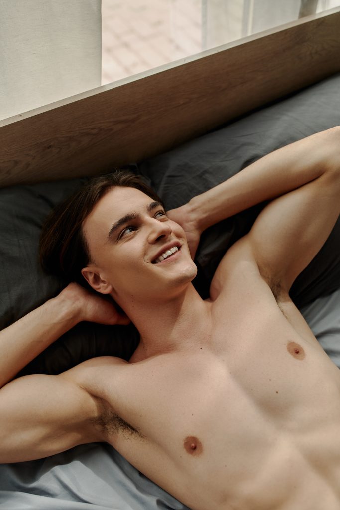 Sensual Male Massage to Enhance Men's Health Happy and attractive shirtless guy smiling and looking content while laying on a bed.