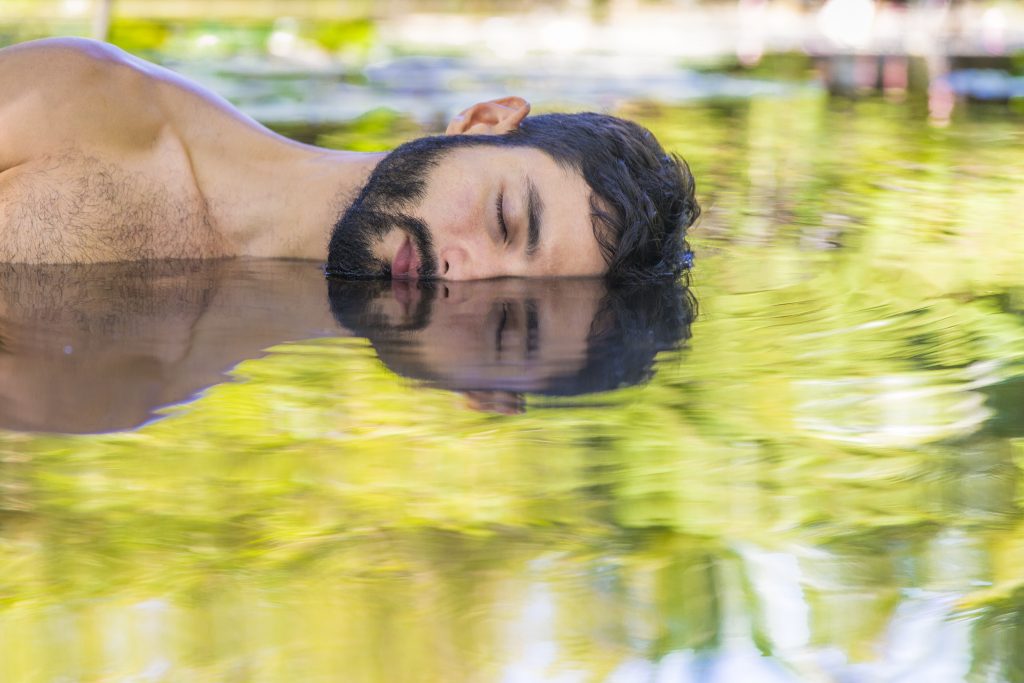 Concept of slowing down and taking time for yourself to ground. A bearded young man laying down on calming water.